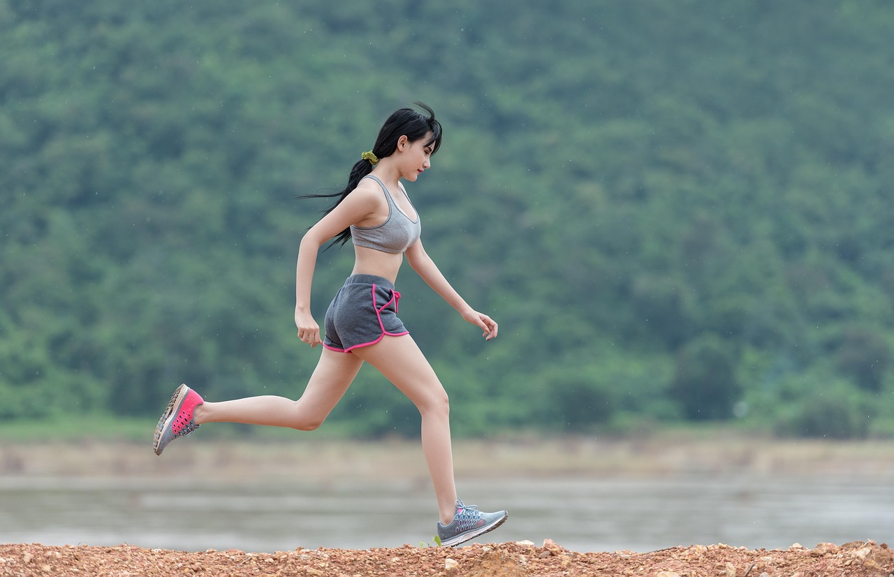 A young woman running on a dirt path near a body of water. She is wearing a gray sports bra and pink shorts, and her hair is tied back in a ponytail. The woman is in mid-stride, with her left leg extended behind her and her right leg bent at the knee. She appears to be focused and determined as she runs. The background is blurred, but it seems to be a wooded area with trees and greenery. The sky is overcast and the overall mood of the image is peaceful and serene.
