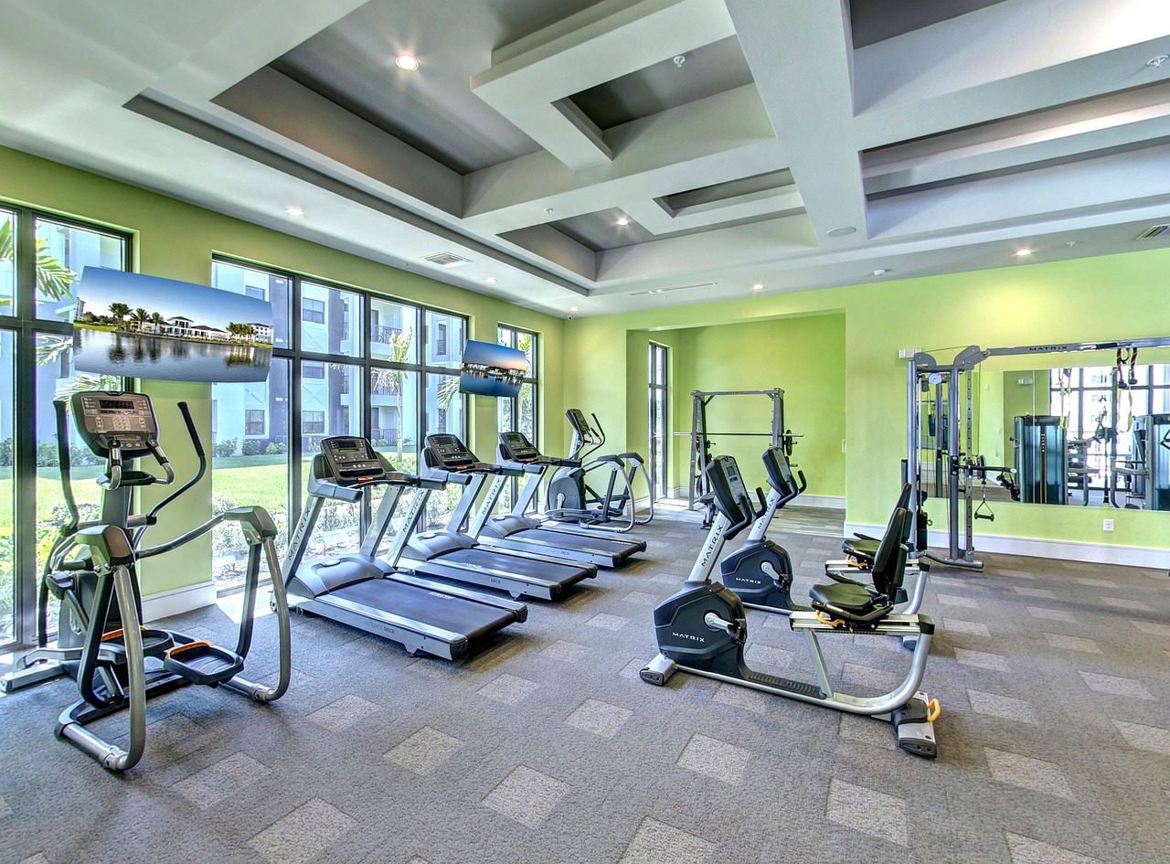 A modern and well-equipped gym with a variety of exercise equipment. There are several treadmills, elliptical machines, and weightlifting machines on the floor. The walls are painted in a bright green color and there are large windows on the left side of the room, allowing natural light to enter. The ceiling is high and has recessed lighting. The floor is covered with a gray carpet. The gym appears to be well-maintained and clean.