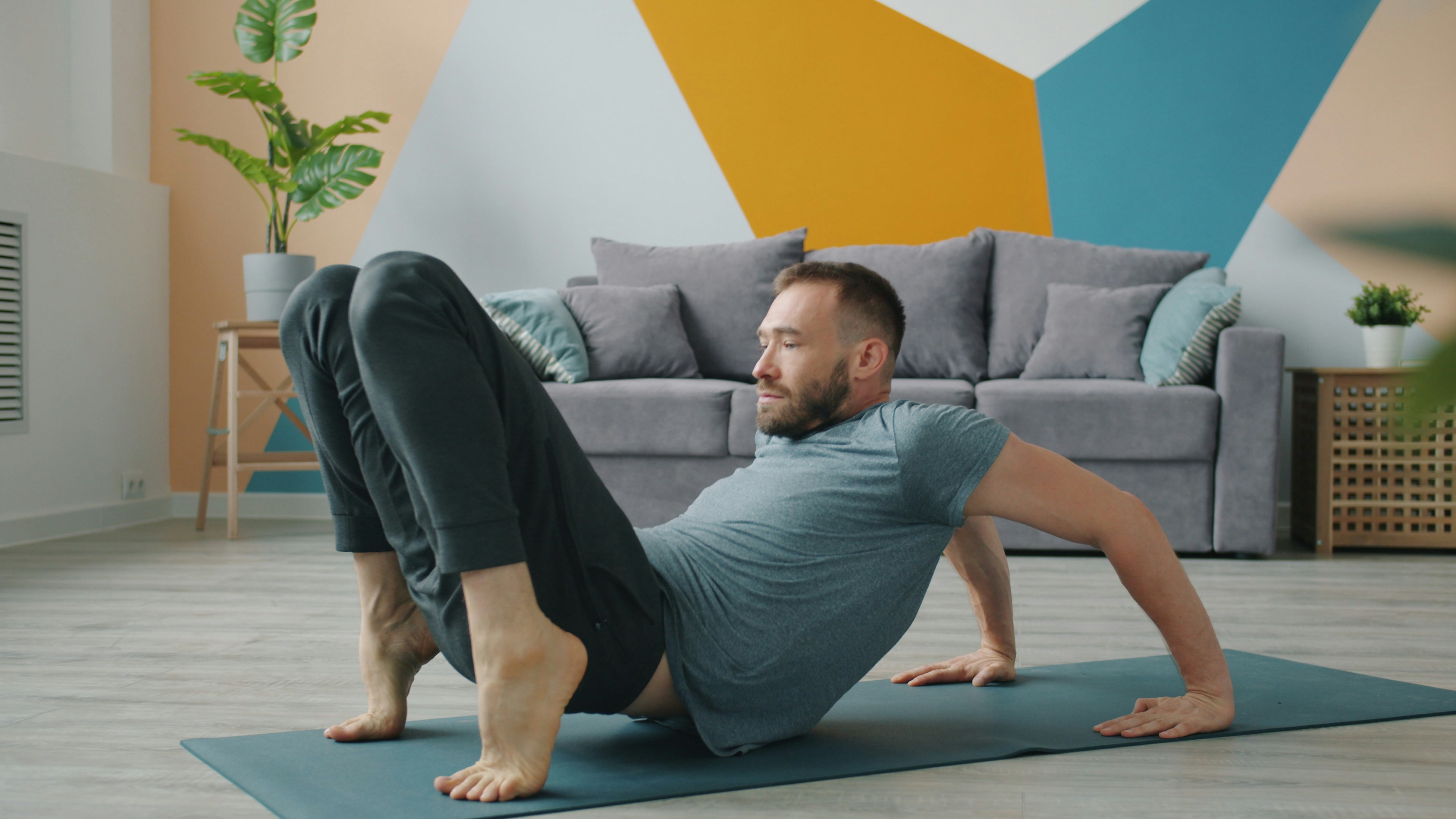 A man performing a yoga pose on a blue mat in a living room. He is wearing a grey t-shirt and black pants. His arms are stret...