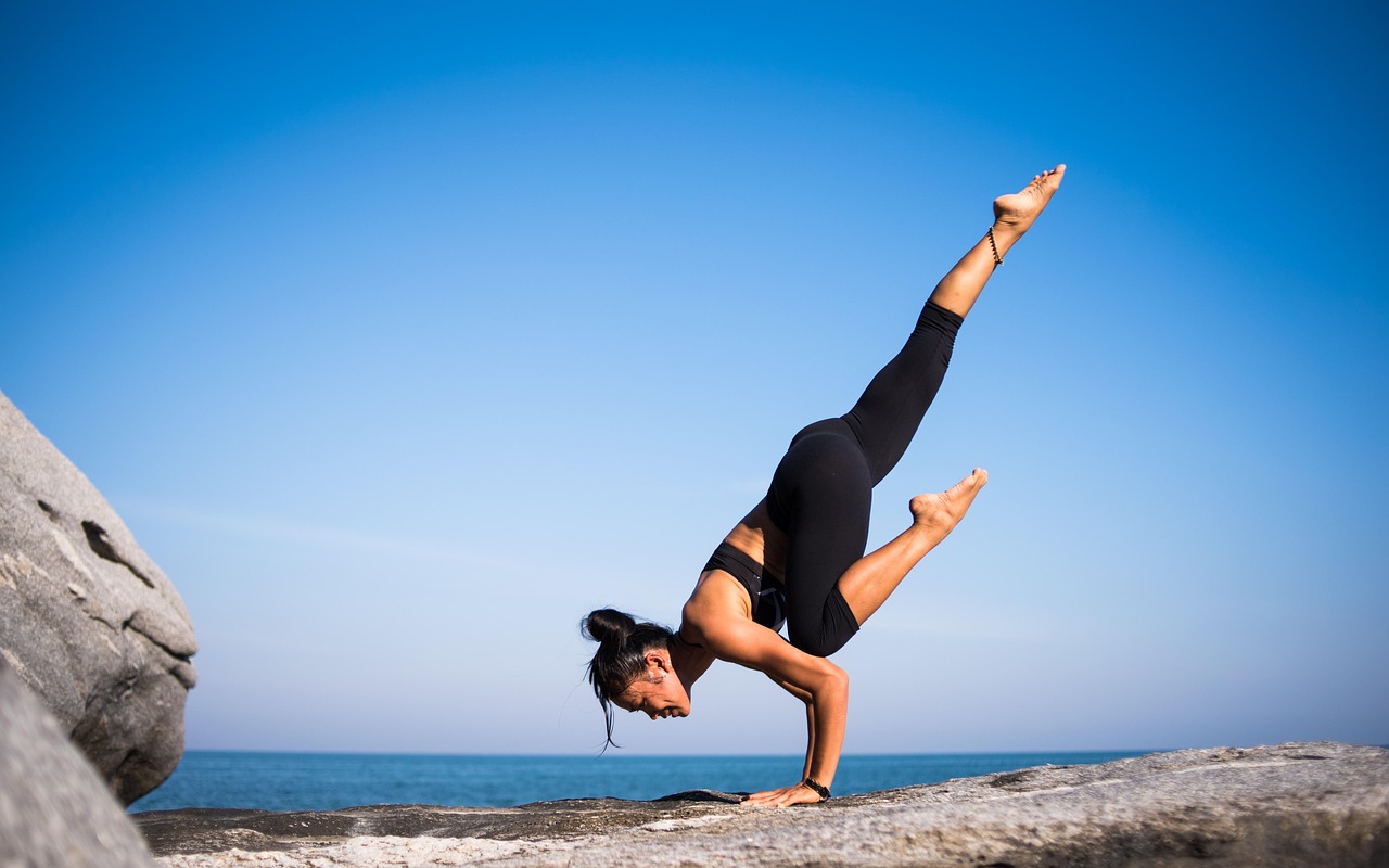 A young woman performing a yoga pose on a rocky cliff overlooking the ocean. She is wearing a black leotard and black legging...