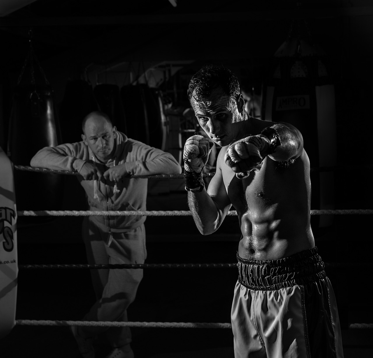 A black and white photograph of a man in a boxing ring. He is shirtless and is wearing boxing gloves and shorts. He appears t...
