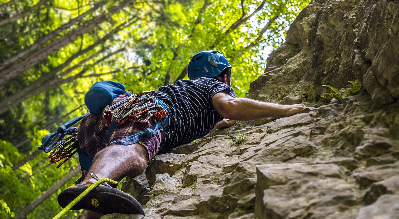 A rock climber scaling a steep cliff in a forest. The climber is wearing a blue helmet, a black and white striped shirt, and ...