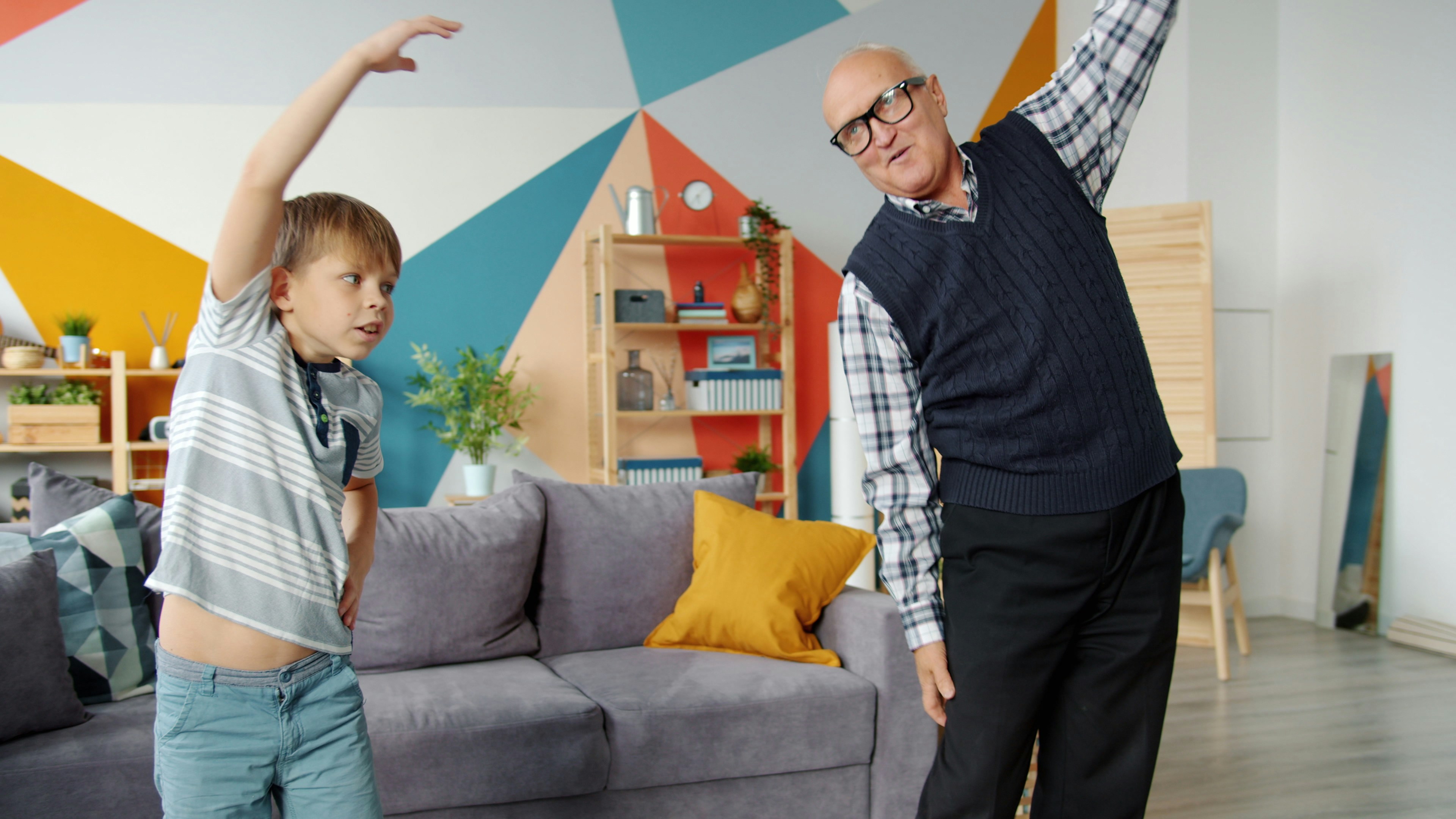 An elderly man and a young boy in a living room. The man is standing on the right side of the image, with his arms stretched ...