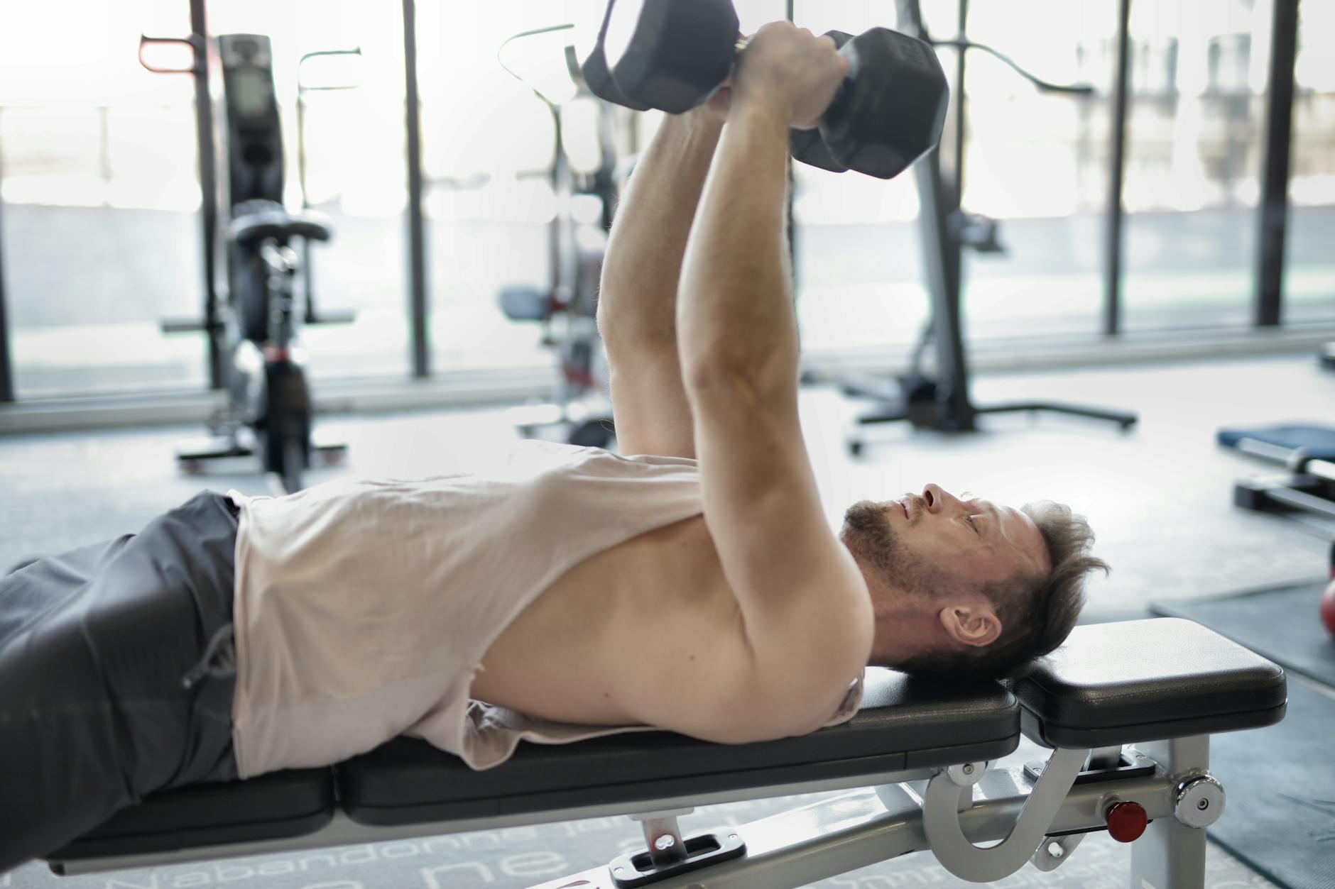 A man lying on a bench in a gym, lifting a pair of dumbbells above his head. He is wearing a white tank top and black shorts....