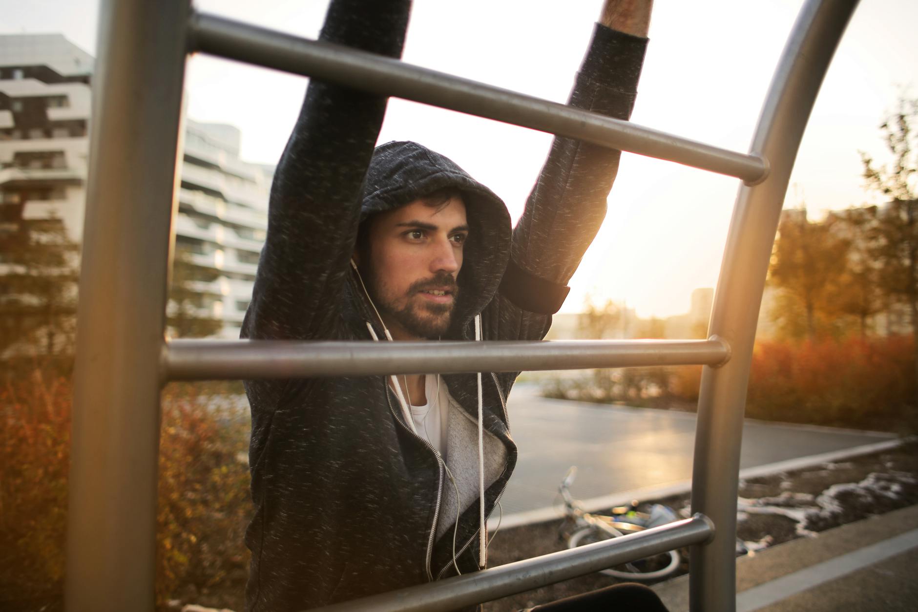 A young man with a beard and mustache, wearing a black hoodie and headphones. He is standing behind a metal railing, with his...
