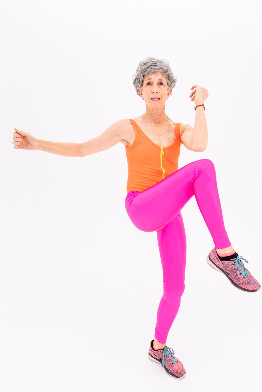 An elderly woman with grey hair, wearing an orange tank top and pink leggings. She is standing on one leg with her left leg b...