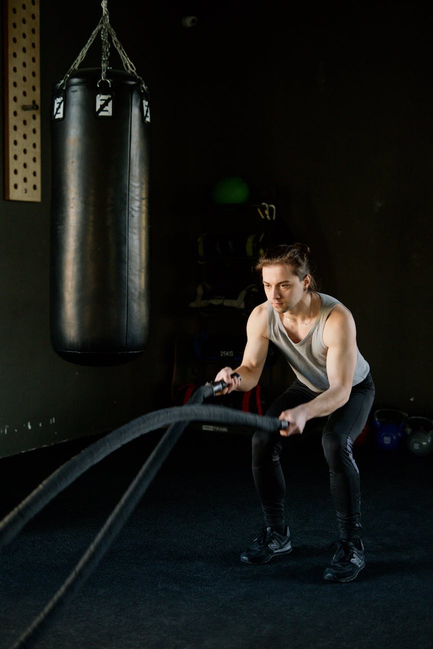A young man in a gym, performing a battle rope exercise. He is wearing a tank top and black leggings, and is holding a black ...