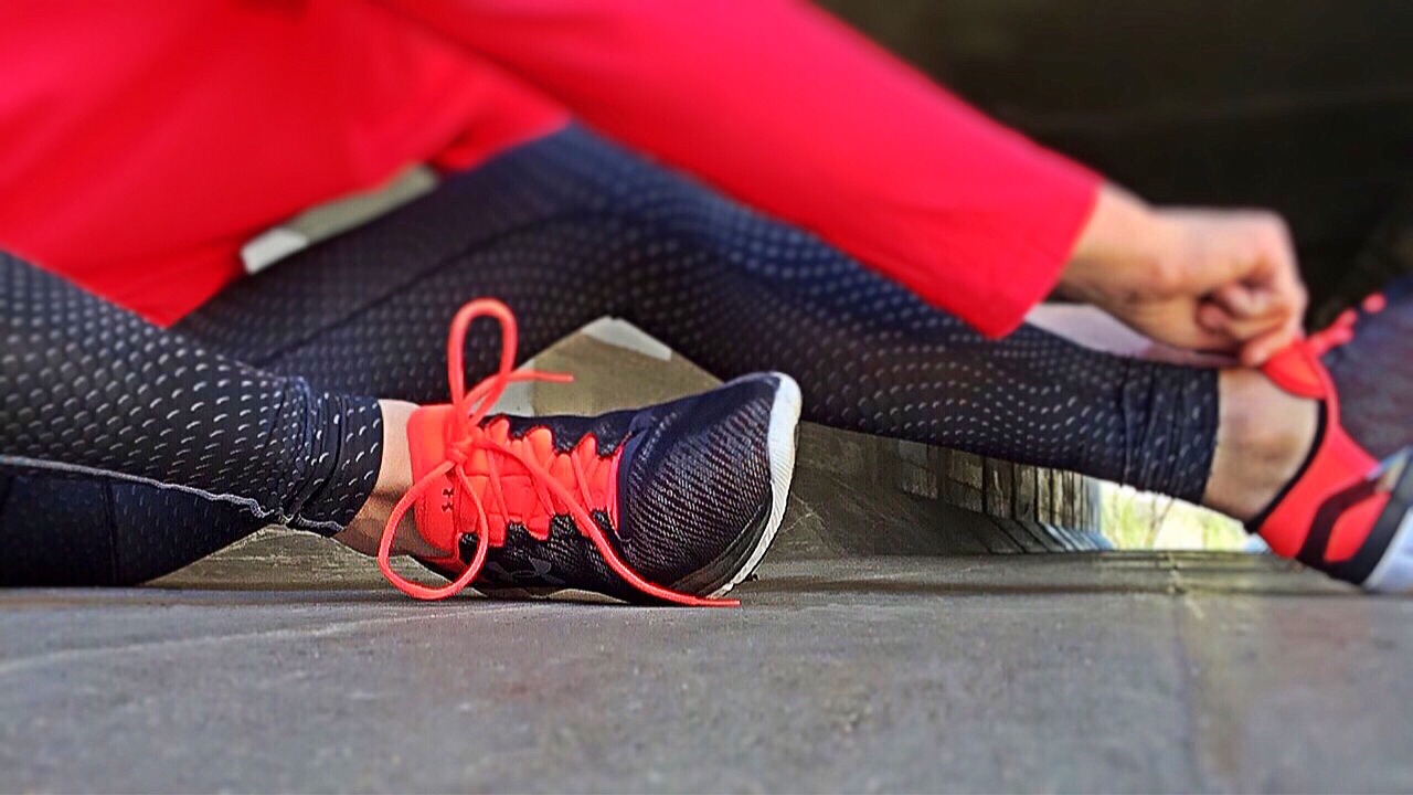 A close-up of a person's legs and feet. The person is wearing red leggings and black sports shoes with red laces. The shoes have a mesh-like pattern on the sides and a black sole. The laces are tied in a bow at the top of the shoe. The background is blurred, but it appears to be an outdoor setting with a concrete floor and a brick wall. The focus of the image is on the person's feet and the shoes.