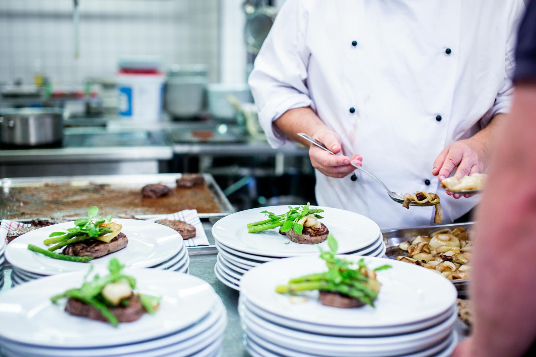 A chef in a kitchen preparing food. He is wearing a white chef's uniform and is holding a pair of tongs in his right hand. In...