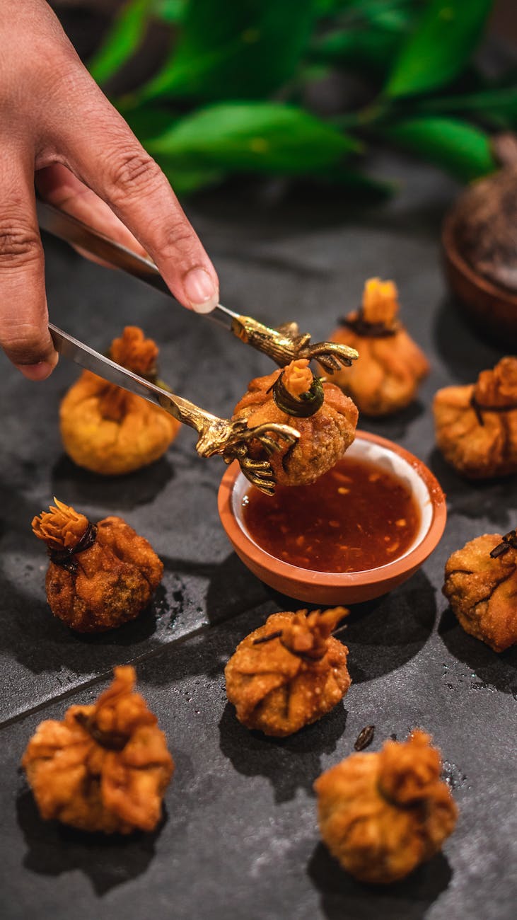 A hand holding a pair of tongs and dipping it into a small bowl of dipping sauce. The tongs are golden brown and appear to be...