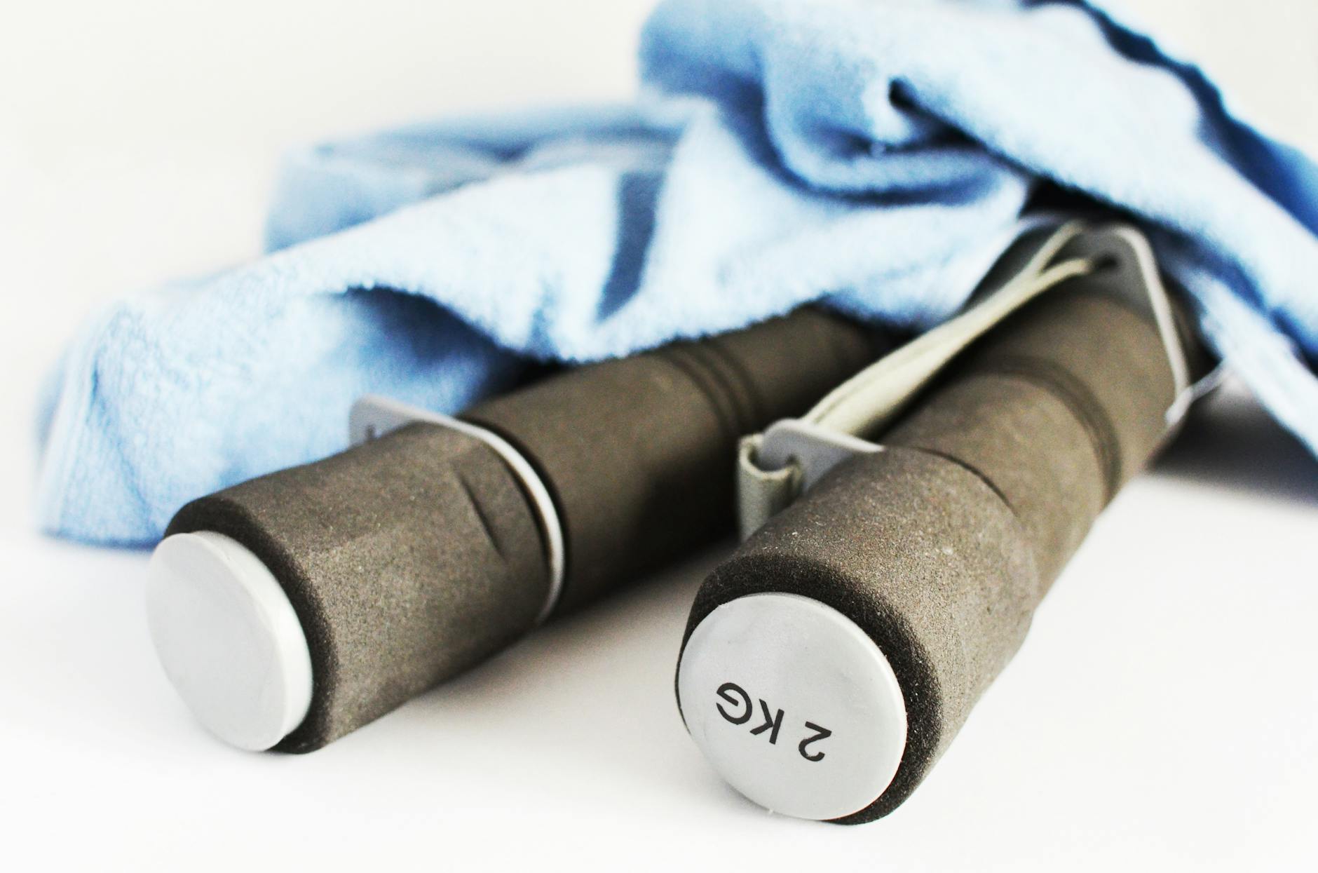 A pair of dumbbells lying on a white surface. The dumbbell on the left is brown in color and has a white handle with the word "2KG" written on it in black letters. The handle is attached to a blue towel, which is folded over the top of the dumbbell. The background is plain white.