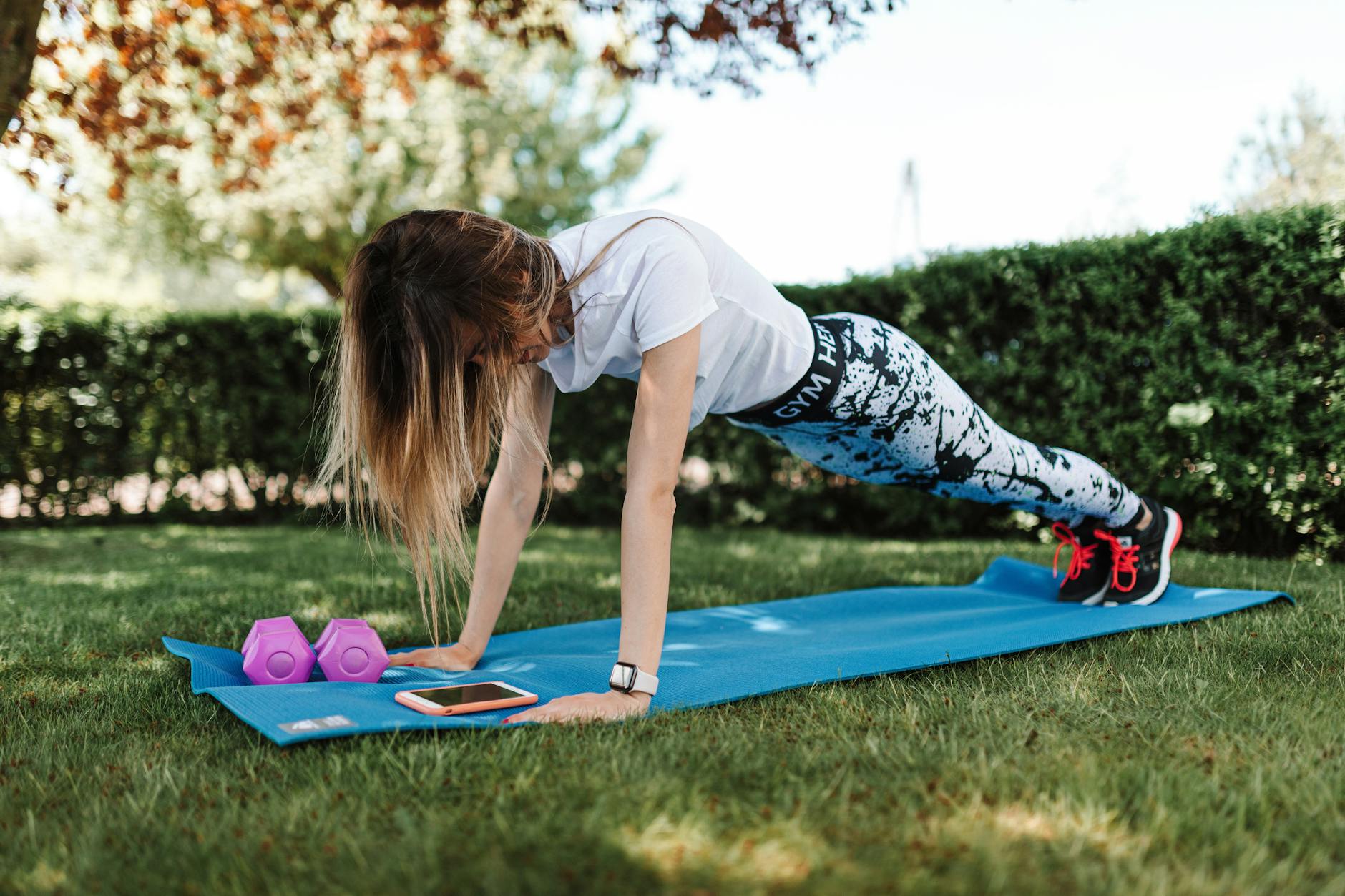 A young woman performing a plank exercise on a blue yoga mat in a garden. She is wearing a white t-shirt, black and white pat...