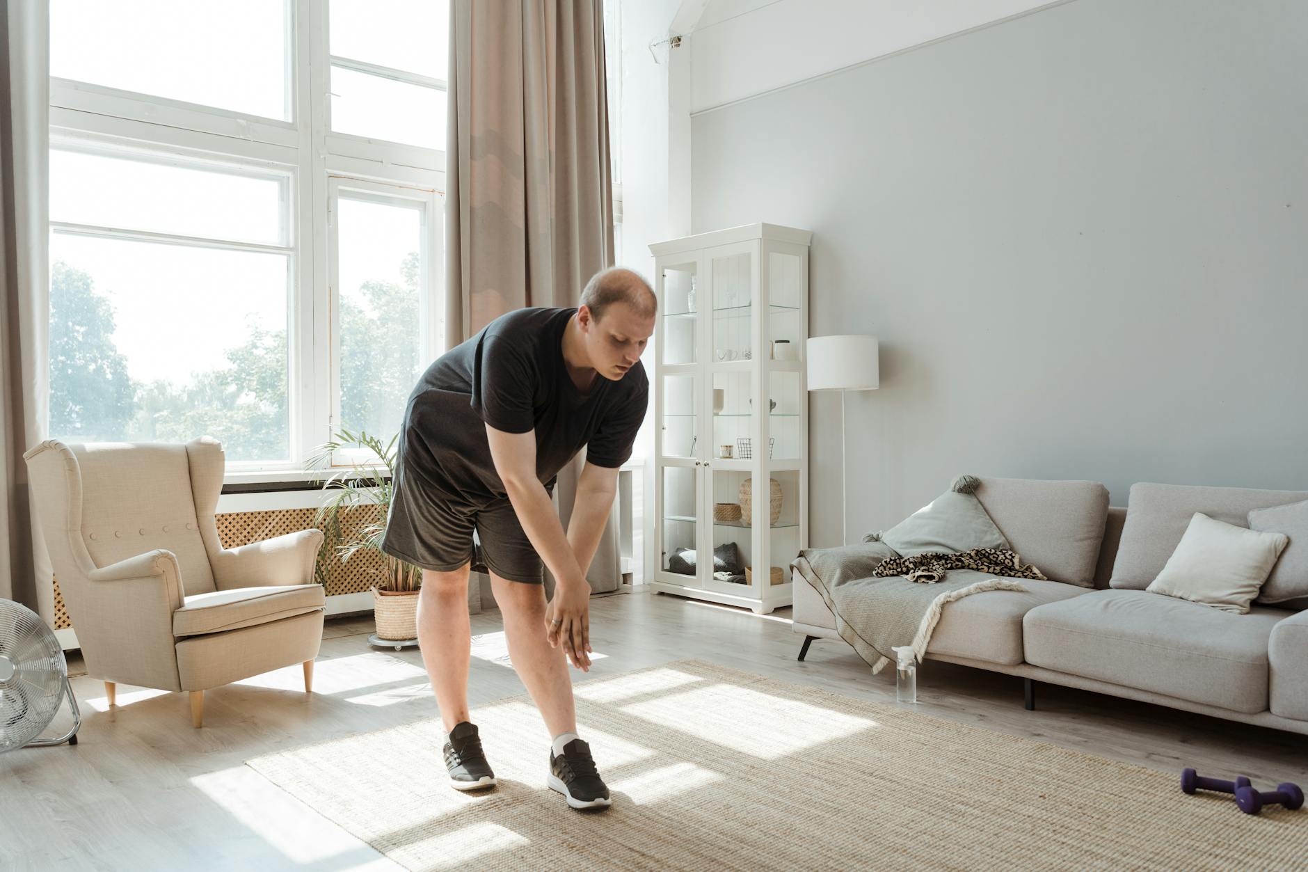 A man in a living room, bending over to pick up something from the floor. He is wearing a black t-shirt, black shorts, and bl...