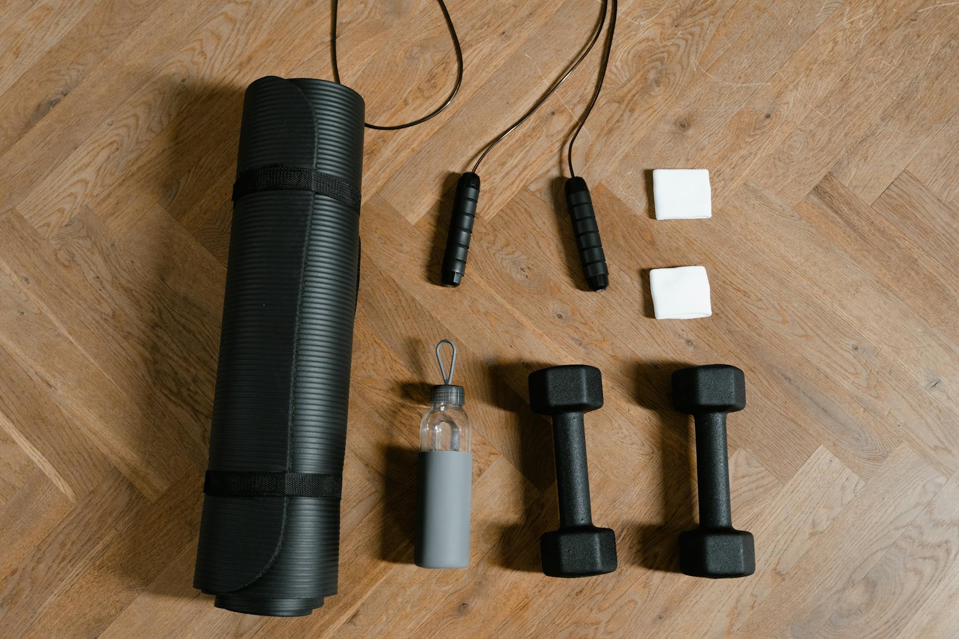 A set of fitness equipment laid out on a wooden floor. On the left side of the image, there is a black yoga mat with a black ...