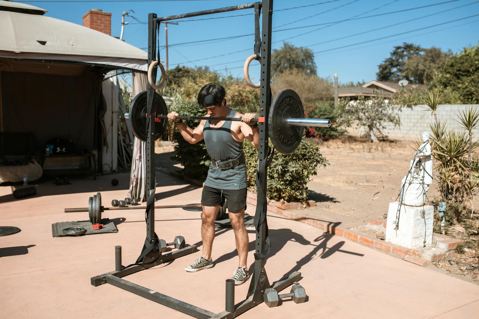 A man lifting a barbell over a weight rack in a backyard. He is wearing a black tank top, shorts, and sneakers. The weight ra...
