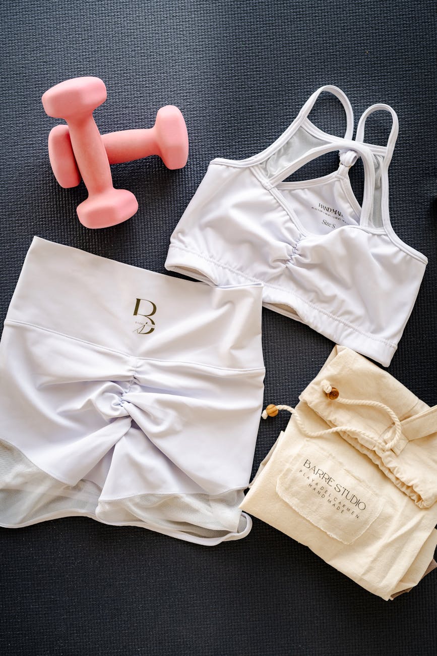 A flat lay of a sports bra, shorts, and a pair of pink dumbbells on a black background. The sports bra is white with thin str...