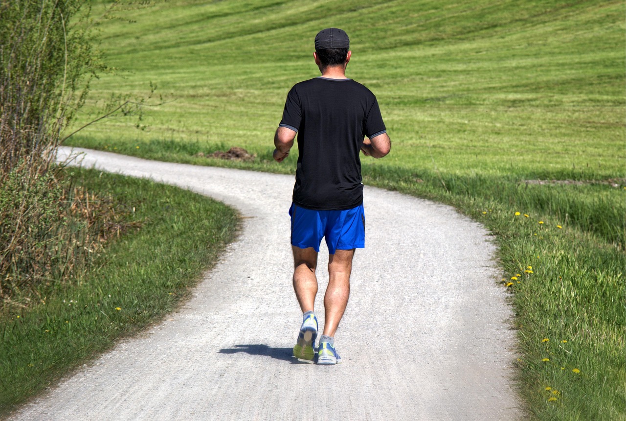 A man walking on a gravel path in a rural area. He is wearing a black t-shirt, blue shorts, and blue sneakers. He has a black...