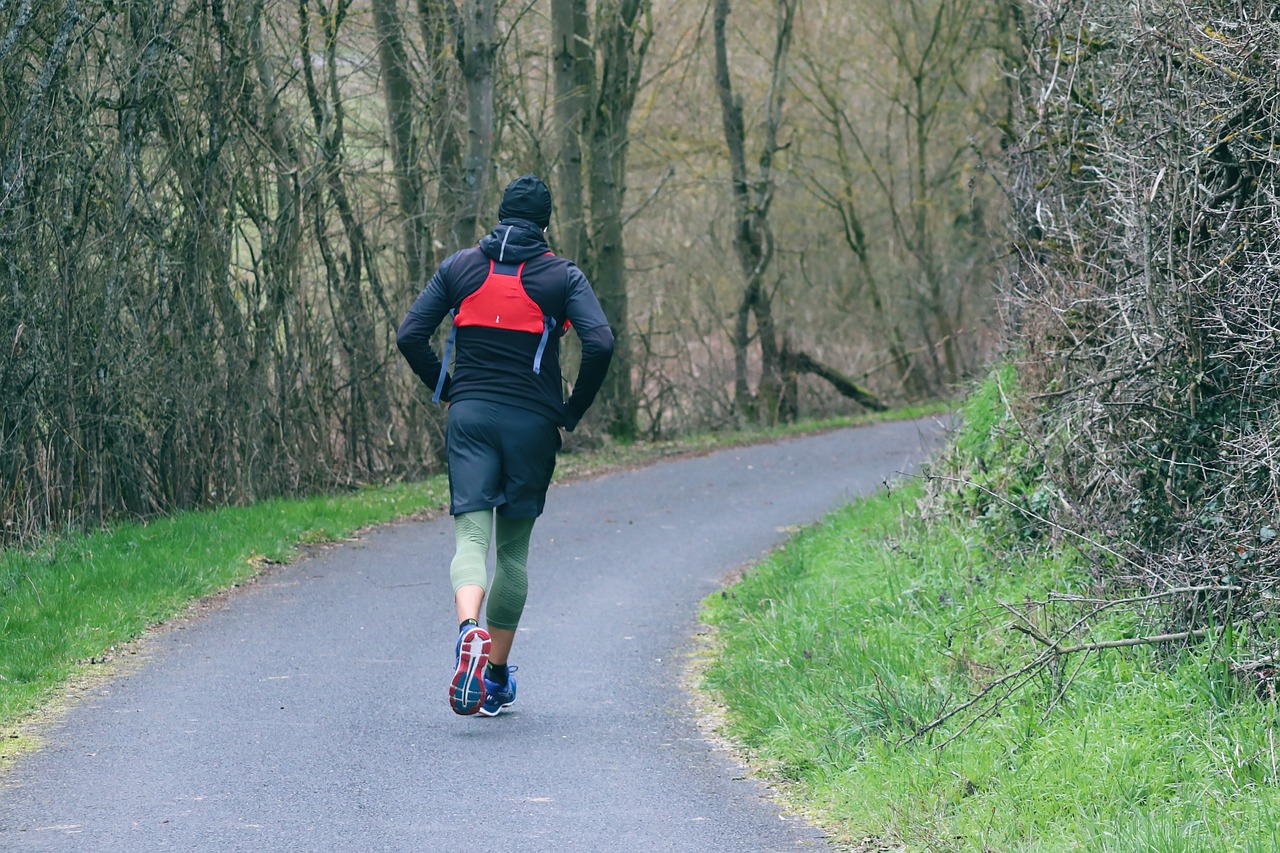 A person running on a paved path in a wooded area. The person is wearing a black jacket, green pants, and red running shoes. ...