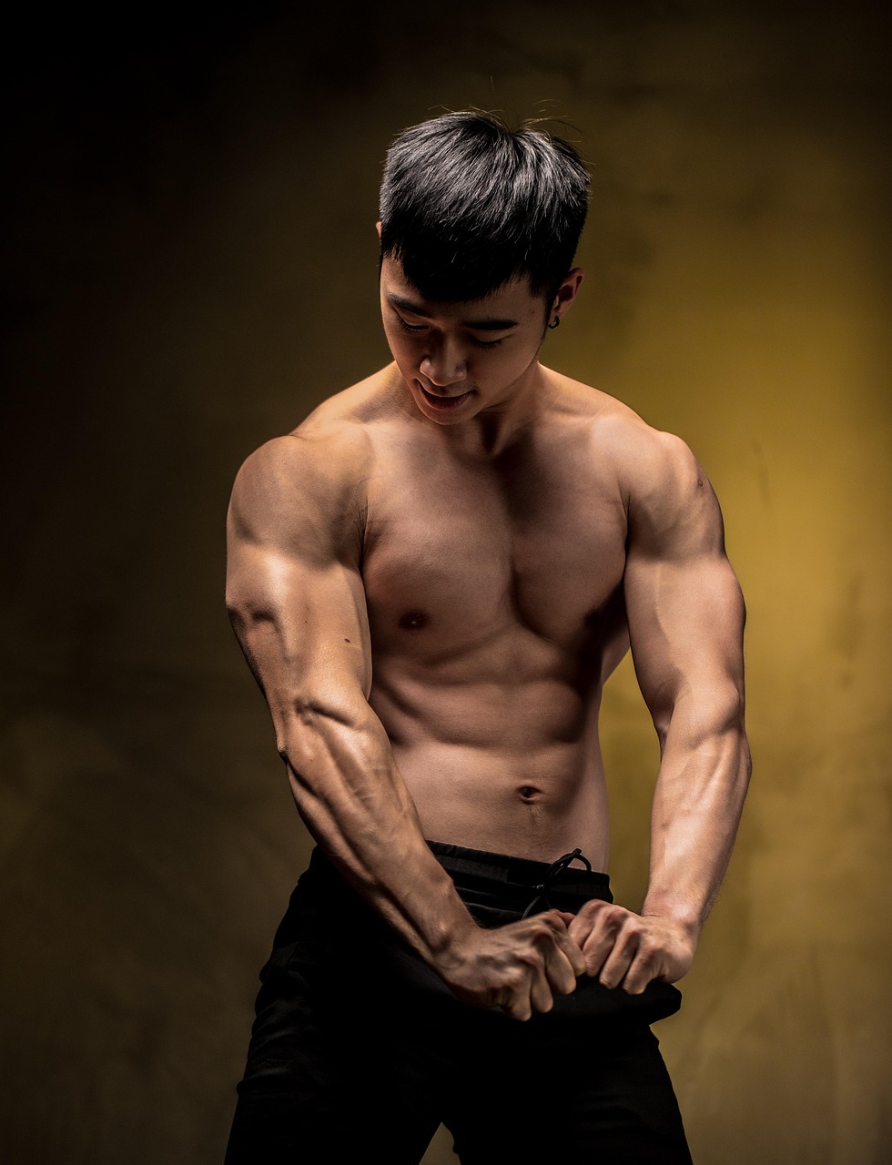 A portrait of a young man with muscular arms and chest. He is shirtless and is wearing black pants. His body is toned and wel...