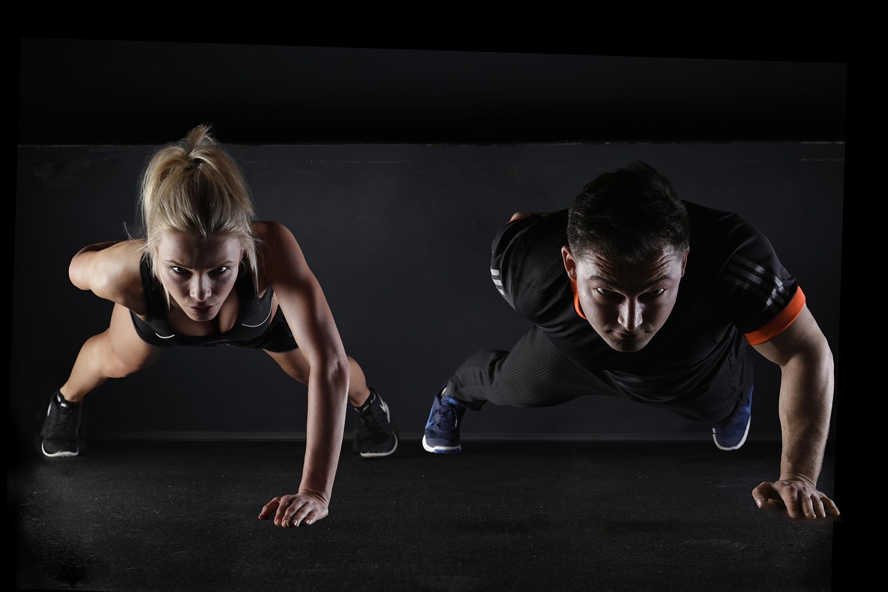 A man and a woman in a push-up position on a black background. The woman is on the left side of the image, with her arms and ...