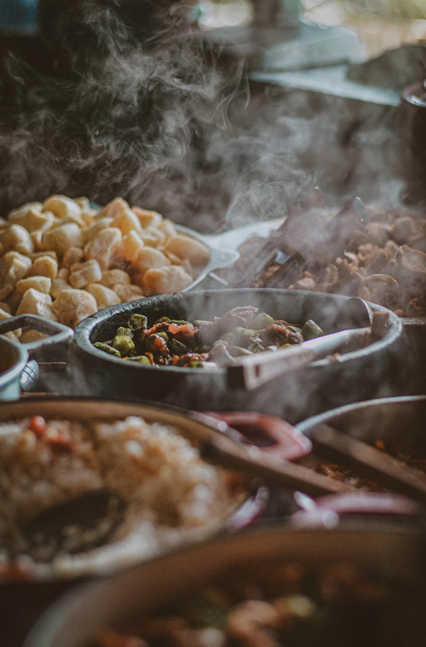 A variety of different types of food being cooked on a grill. There are several large pots and pans filled with different dis...