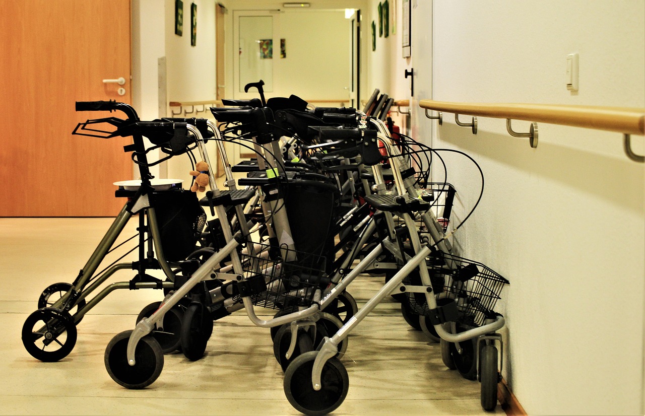 A row of walkers lined up against a white wall in a hallway. The walkers are made of metal and have black handles and wheels....