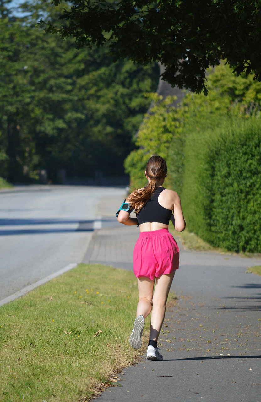 A young woman jogging on a paved path in a park. She is wearing a black sports bra and pink shorts, and white sneakers. Her h...