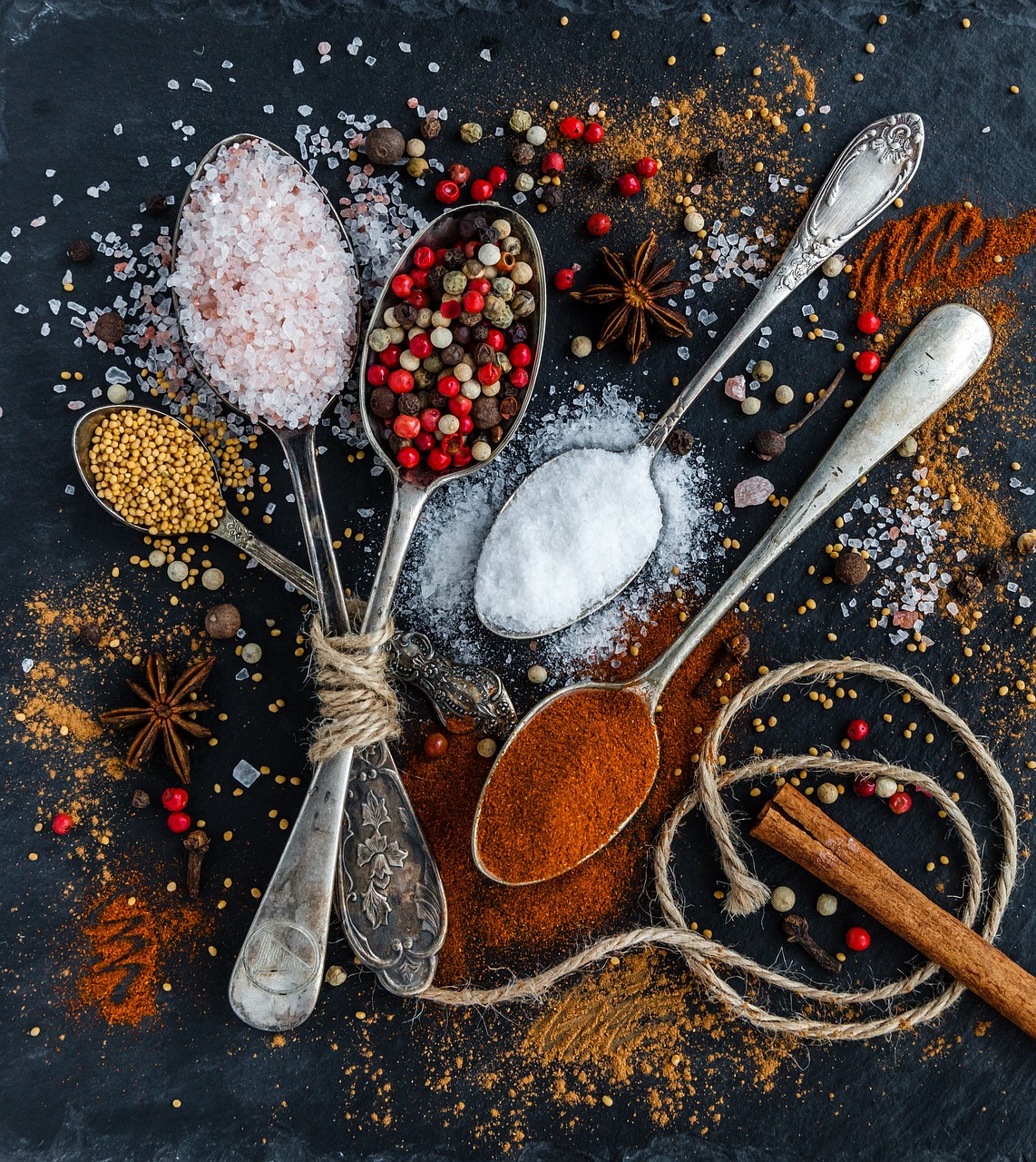 A flat lay of various spices and herbs arranged on a dark grey background. There are four silver spoons in the image, each wi...