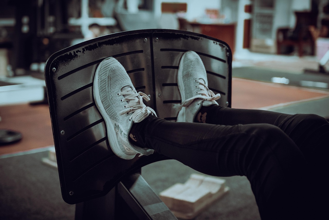 A person's feet resting on a black chair in a gym. The person is wearing black leggings and white sneakers with white laces. ...